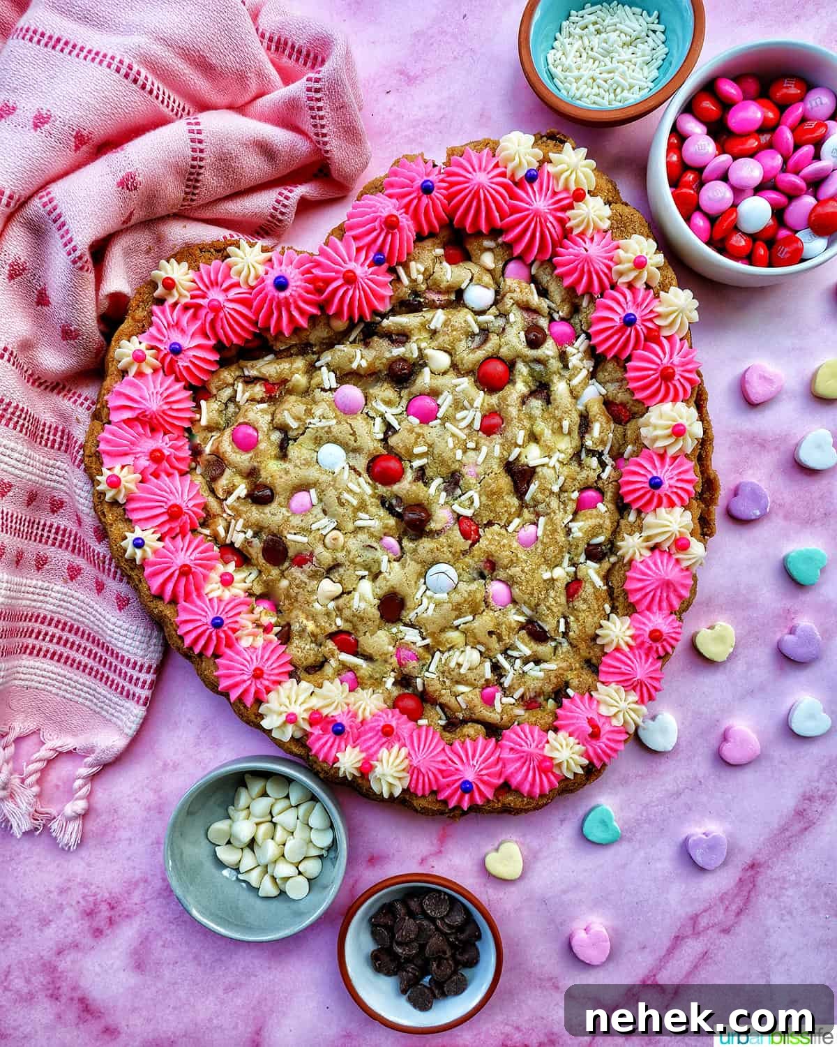 A Valentine's Day Cookie Cake with pink frosting, pink, red and white M & M candies, conversation hearts candies sprinkled around the cookie cake, and white sprinkles.