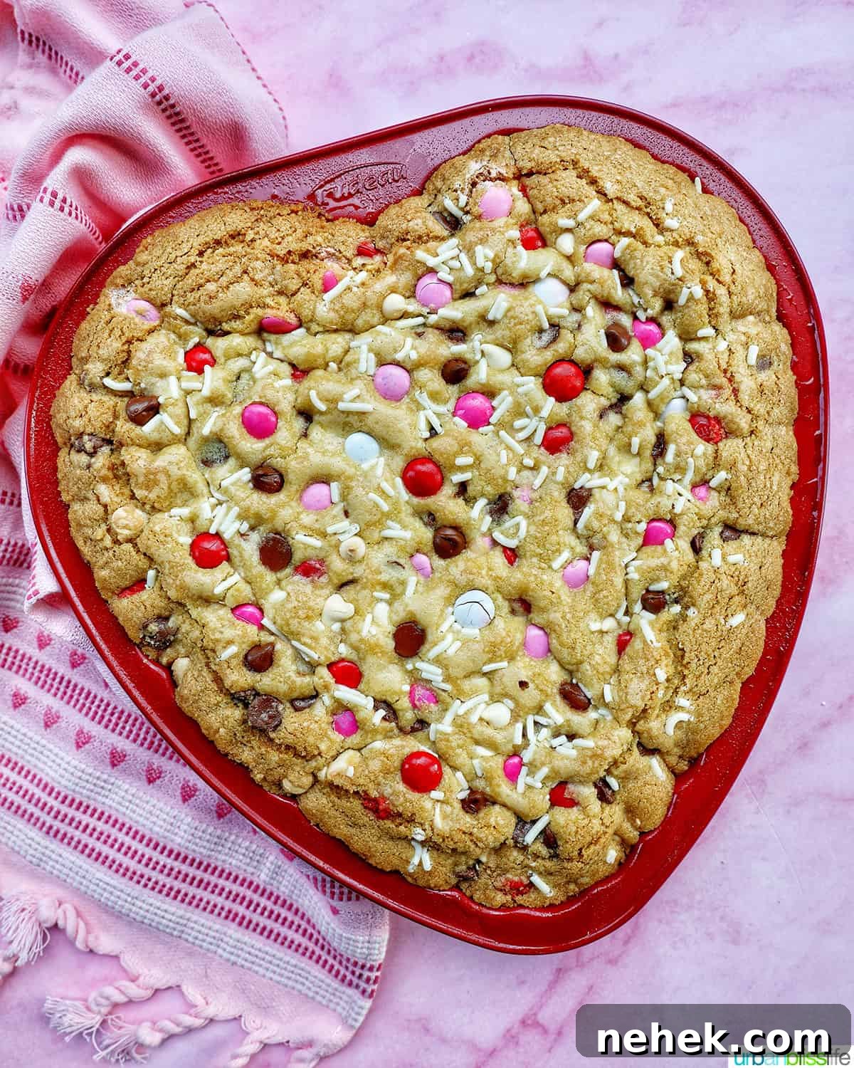 a heart-shaped cookie cake baked in a red pan on pink background.