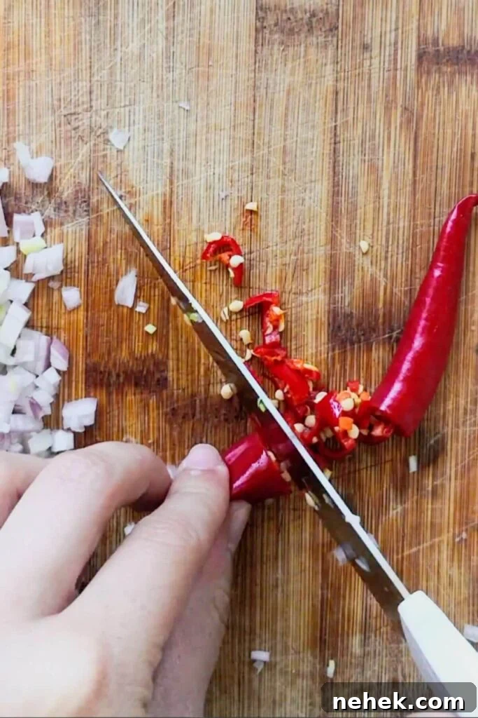 Hand with knife chopping red chilli on a wooden board.