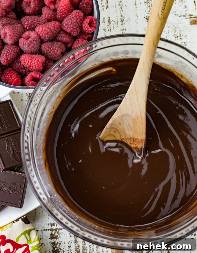 A mixing bowl of chocolate ganache with a wooden spoon. A bowl of fresh raspberries and bars of chocolate next to the mixing bowl.