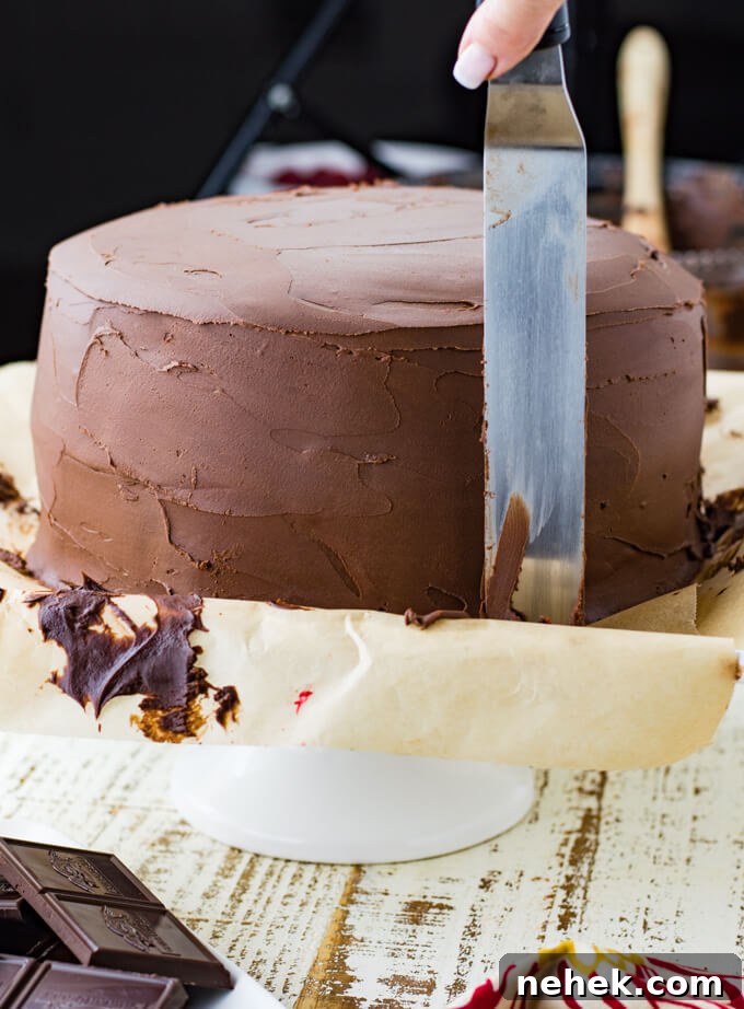 A layered chocolate cake sitting on parchment paper that is on a cake plate. A cake spatula is used to frost the cake.