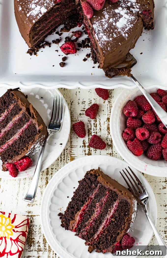 Two pieces of 4 layered chocolate cake with a raspberry filling. The cake sits on a cake plate with slices taken out. A bowl of red raspberries sit next to the cake.