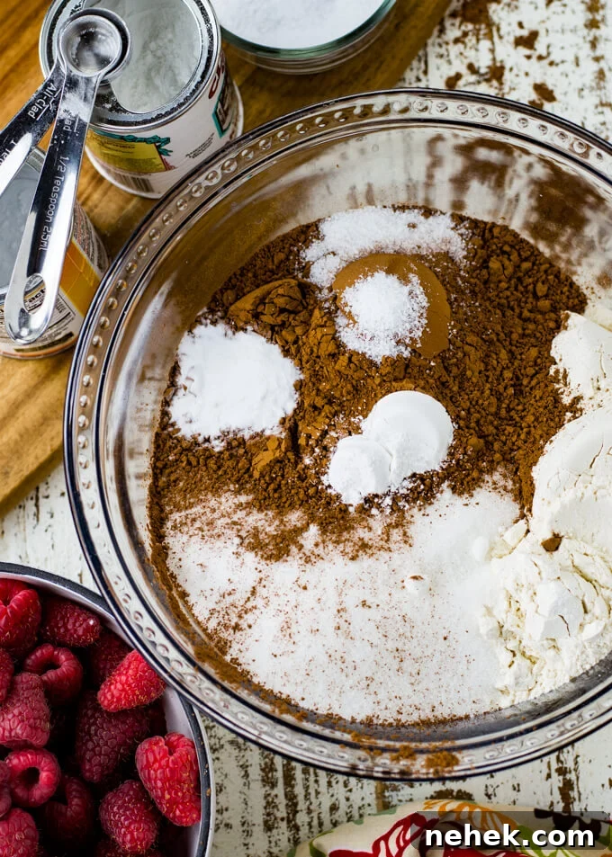 A clear mixing bowl with dry ingredients for a layered chocolate cake. Some of the ingredients sit next to the mixing bowl. A bowl of fresh raspberries are also in the photo.