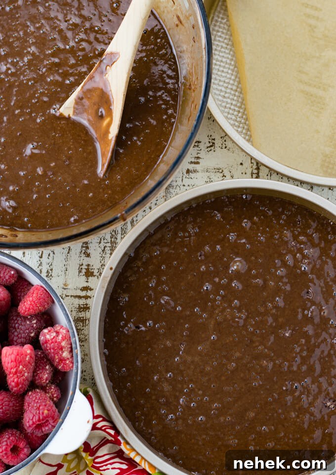 A pan of chocolate cake batter and a bowl of the same batter with a wooden spoon. There's another pan with a piece of parchment paper in it. A bowl fo fresh raspberries sit next to the cake batter.