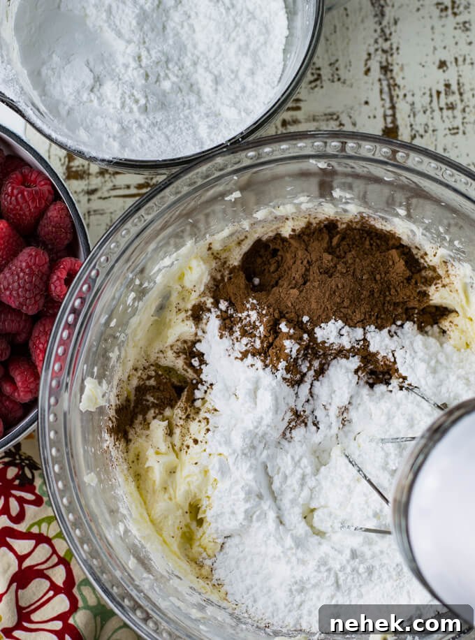 A mixing bowl with ingredients for chocolate icing: powdered sugar, butter, cream cheese, and cocoa. Bowls of powdered sugar and fresh raspberries sit next to the bowl.