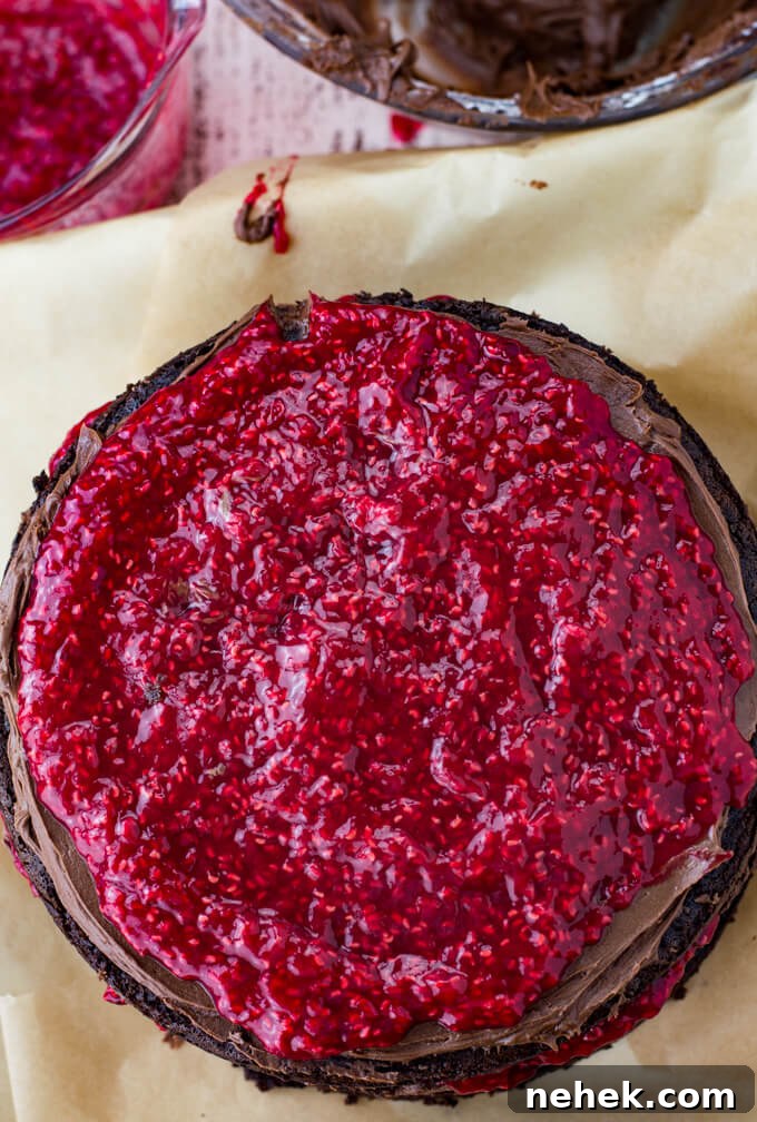 A raspberry filling on a layer of chocolate cake. A bowl of frosting and a bowl of raspberry filling sit next to the cake.