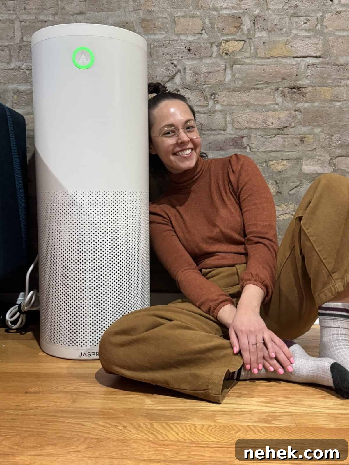 Smiling woman sitting on the floor, leaning against a Jaspr air purifier, with a cozy brick wall background.