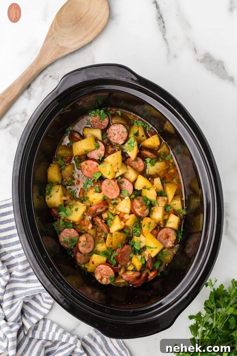 a prep image of a crockpot full of cubed potatoes, sausage, onion, red bell pepper, green bell pepper, garlic, and spices with a wooden spoon next to the crock pot and parsley.