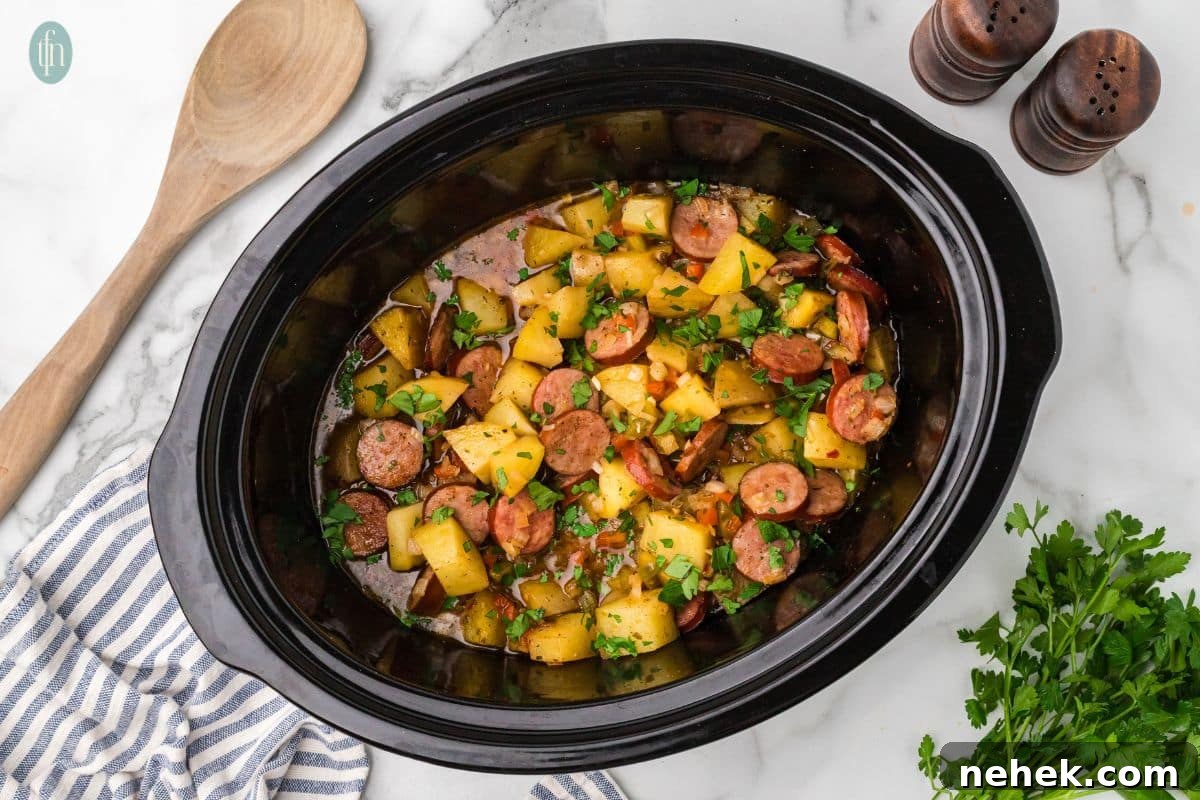 a horizontal image of sausages and potatoes in slow cooker in a crock pot with salt and pepper shakers, a wooden spoon and parsley next to it.