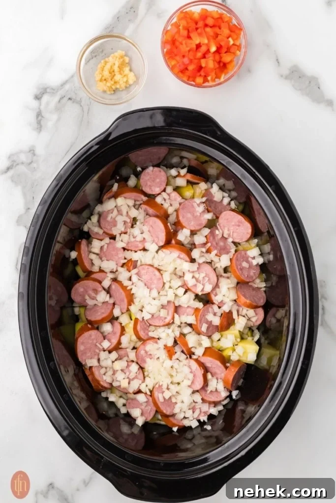 a prep image of a crockpot full of cubed potatoes, chopped onion, and sausage with a bowl of red bell pepper and garlic in bowls next to it.