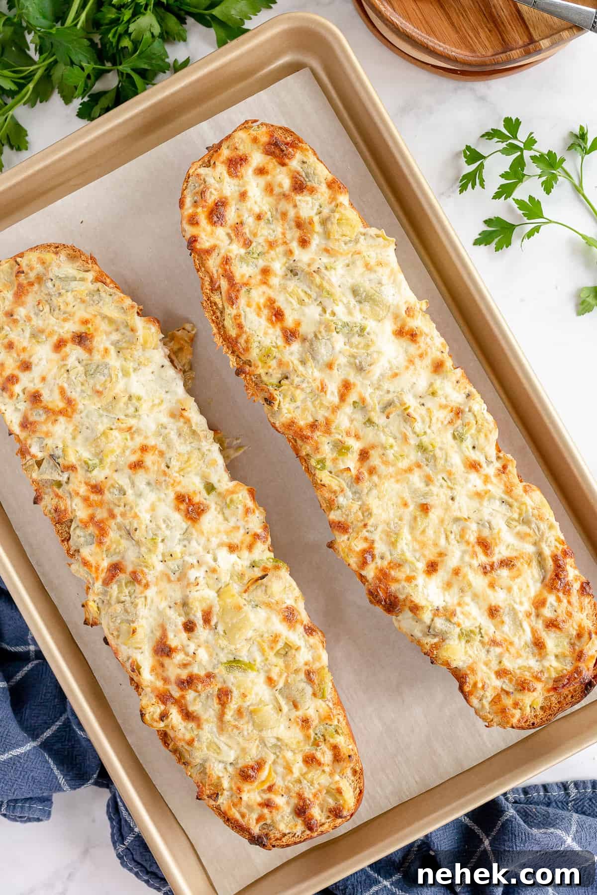Two halves of a loaf of French bread topped with a browned cheese artichoke mixture on a baking sheet.