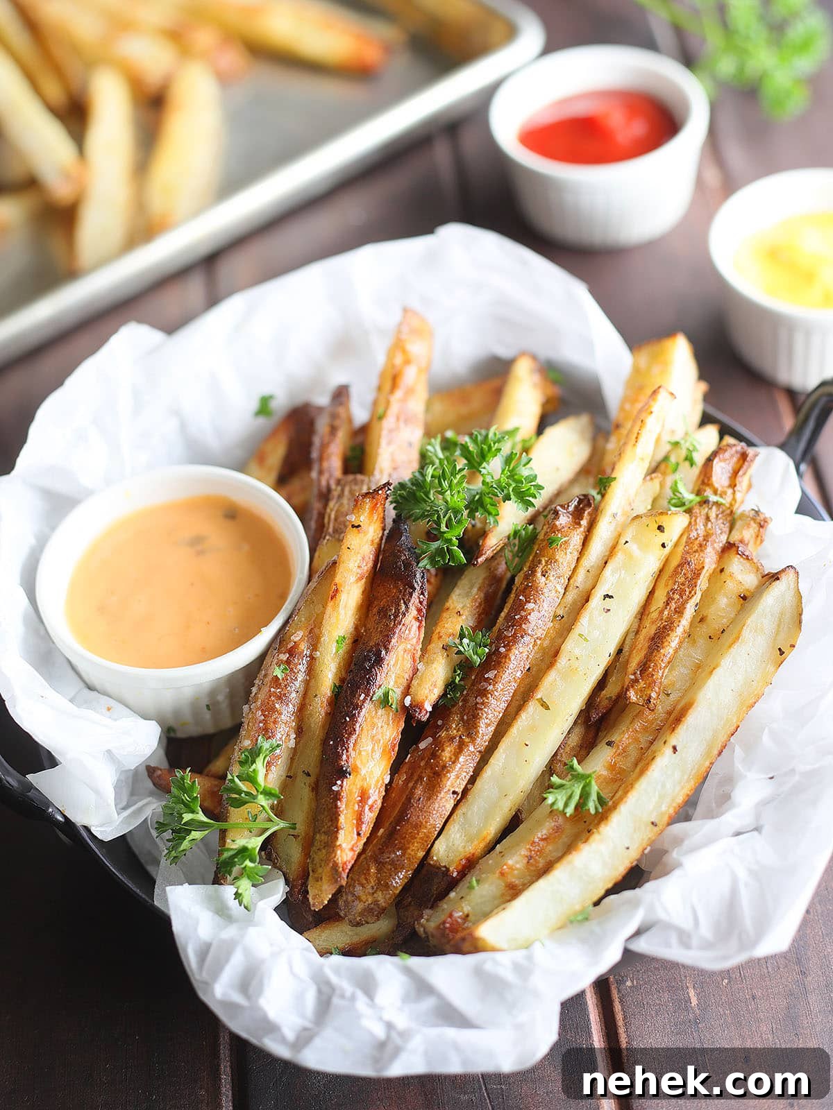 Crispy Oven-Baked French Fries Recipe for Perfect Crunch 9 Basket of oven baked French fries garnished with salt and chopped fresh parsley with dipping sauces to the side.
