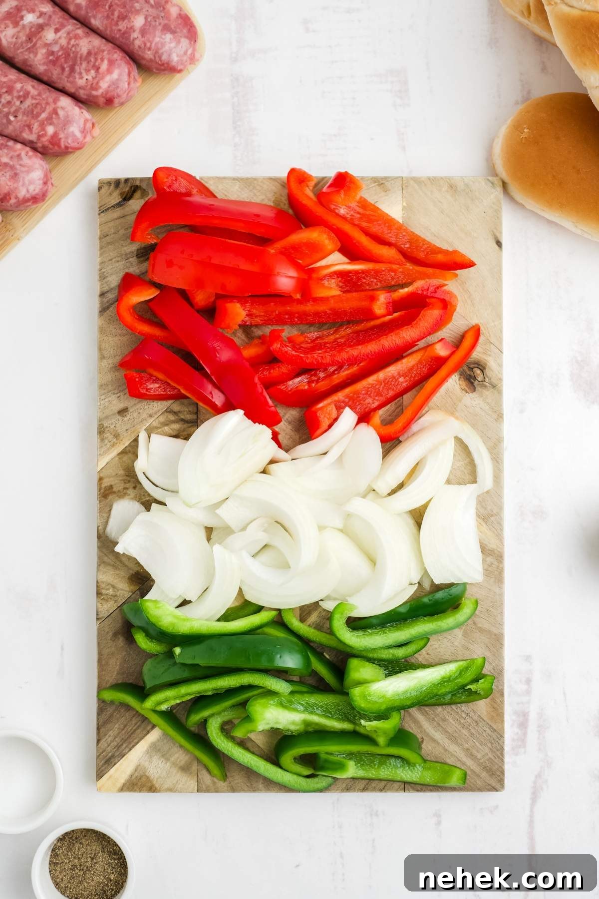 Sliced red bell peppers, white onions, and green bell peppers displayed on a wooden cutting board, with raw sausages and bread buns visible in the background—perfect ingredients for delicious air fryer brats with peppers and onions.
