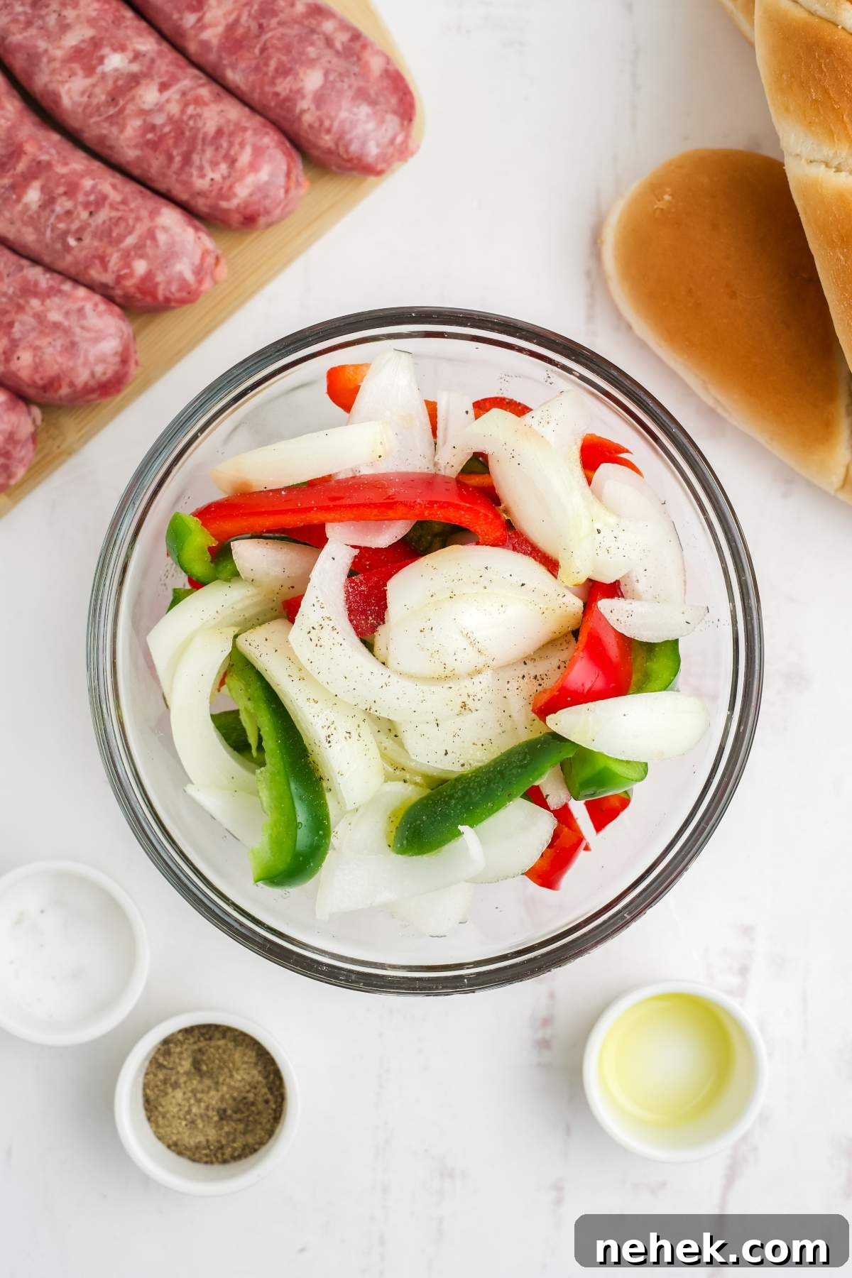 A bowl of sliced red and green bell peppers and onions, seasoned with black pepper. Surrounding the bowl are air fryer brats with peppers and onions, bread rolls, and small containers of salt, pepper, and oil.