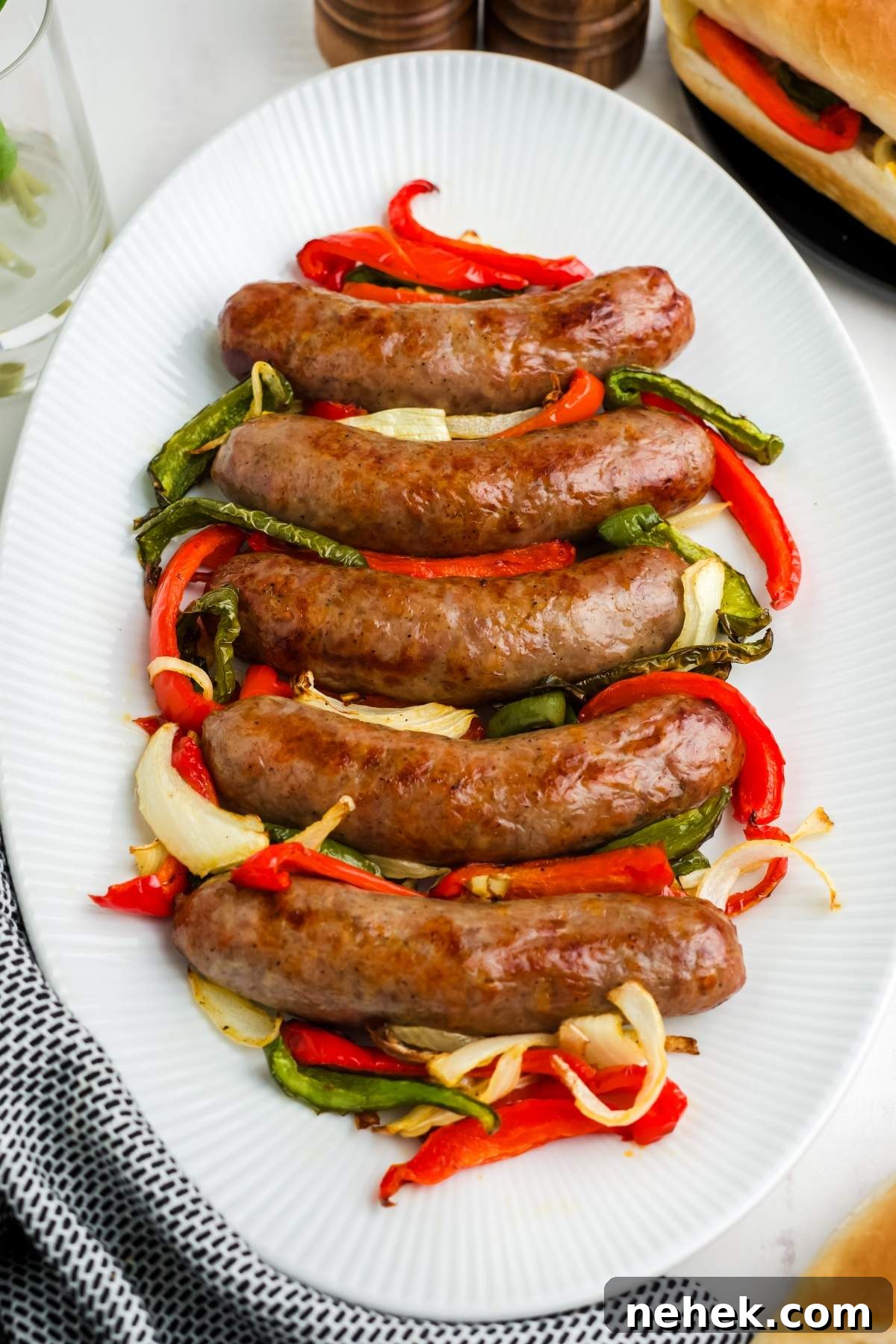 A white oval platter showcases air fryer brats with peppers and onions, featuring five perfectly cooked sausages nestled on a vibrant mix of red and green peppers as well as onions.