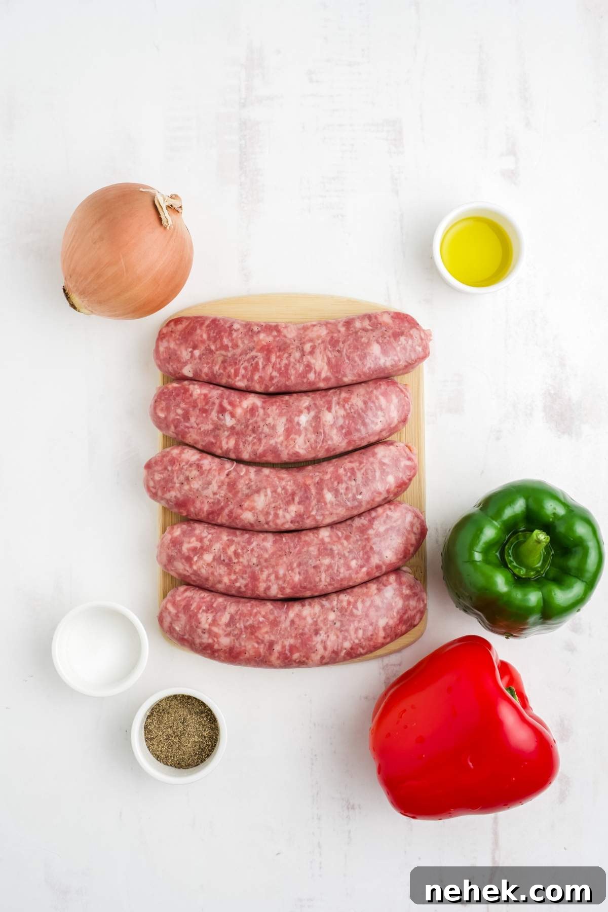 Top-down view of five raw brats on a wooden board, surrounded by onions, green and red bell peppers, a bowl of oil, salt, and pepper on a white surface.