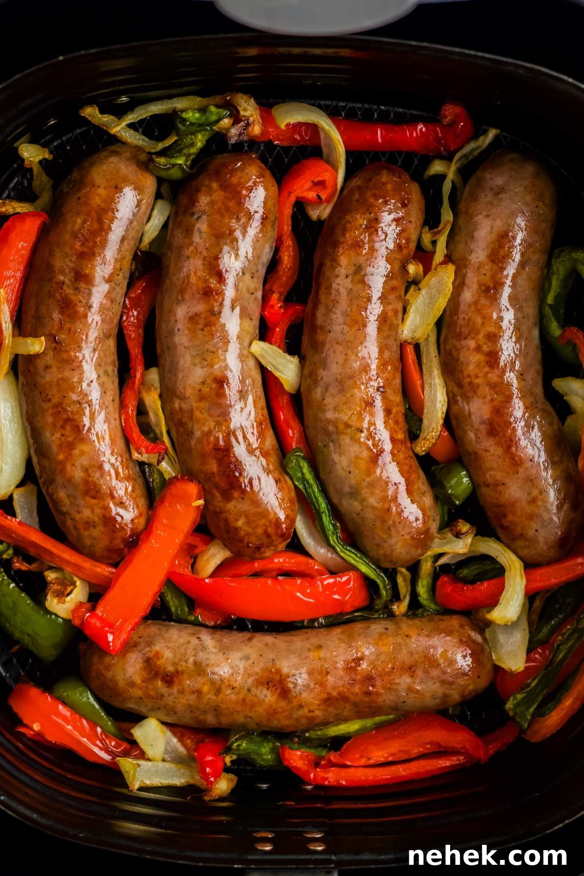 Five cooked brats with peppers and onions atop diced bell peppers and onions in an air fryer basket.