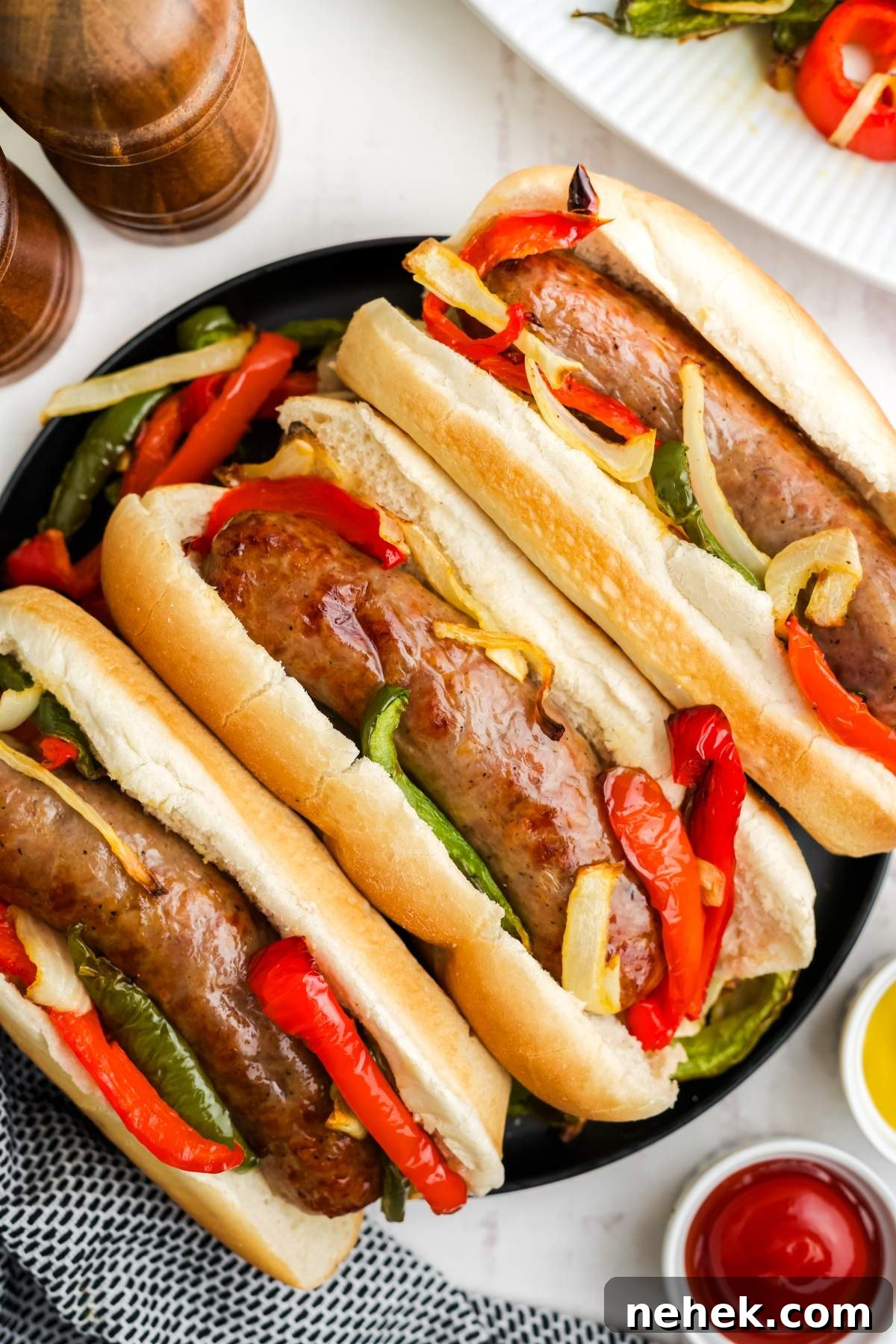 Three air fried brats in buns, topped with sautéed red and green bell peppers and onions, arranged on a black plate. Condiment bottles and a pepper grinder are partially visible in the background.