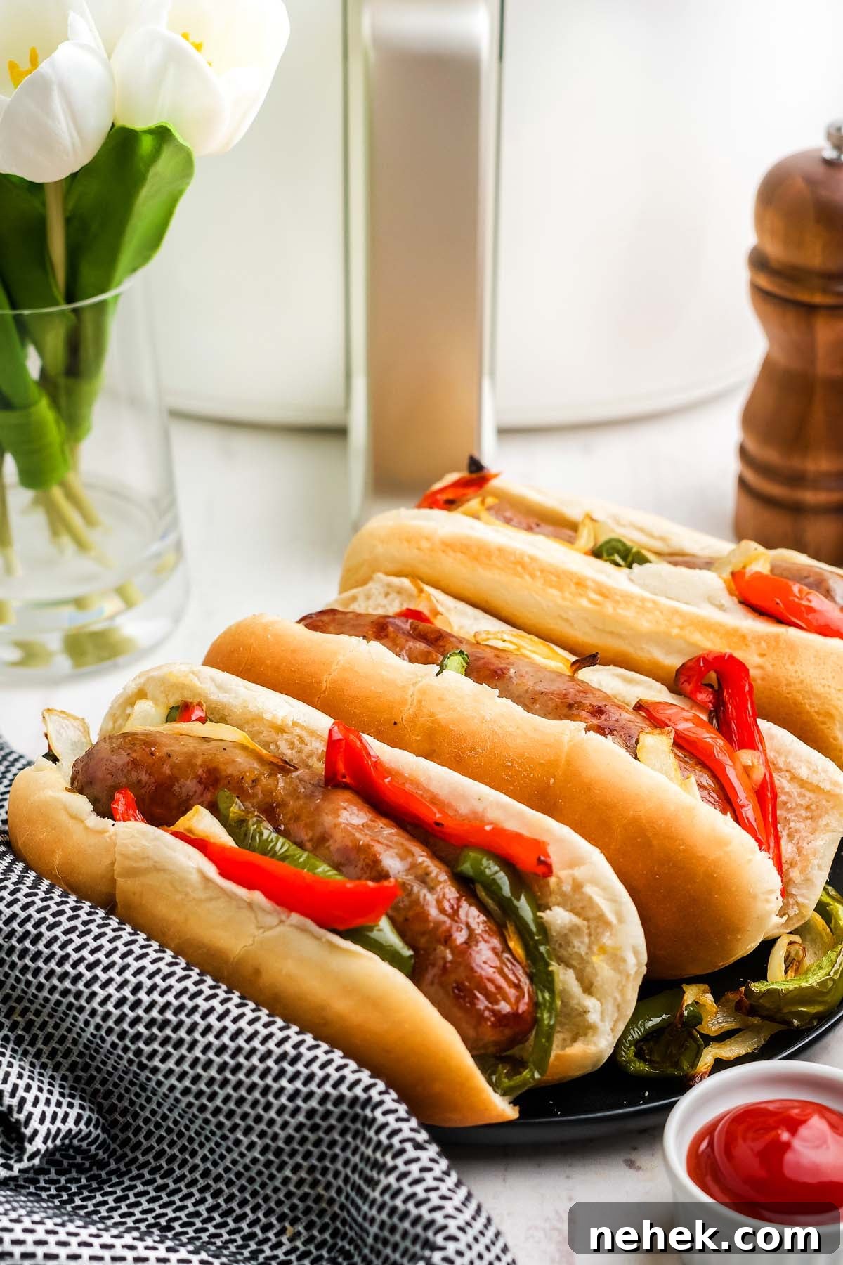 A plate of three air fried bratwurst sausage with sautéed peppers and onions is placed on a checked napkin. Nearby are a bowl of ketchup, a pepper mill, a milk bottle, and a vase with white tulips.