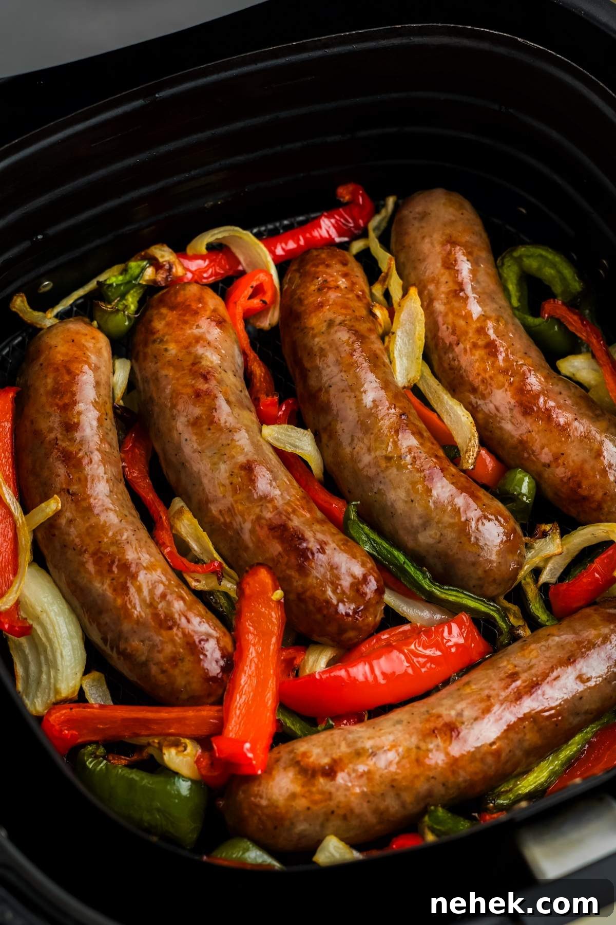 Five cooked brats lie on a bed of sliced red and green bell peppers and onions fresh from the air fryer.