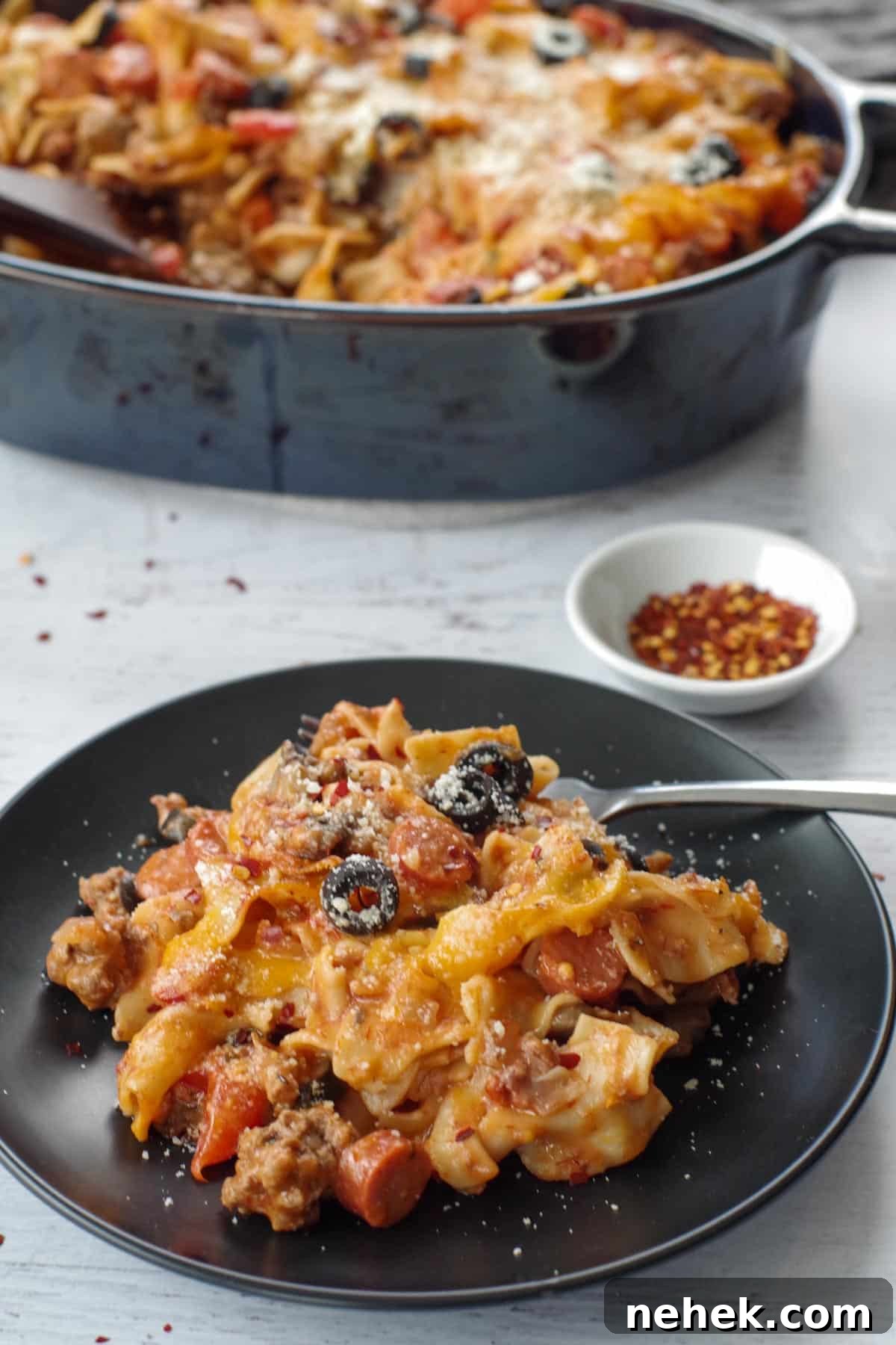 Low-Carb Pizza Casserole with Veggies and Mozzarella 12 single serving of healthy pizza casserole on a black plate with fork and dish of crushed red pepper flakes, with whole casserole in background