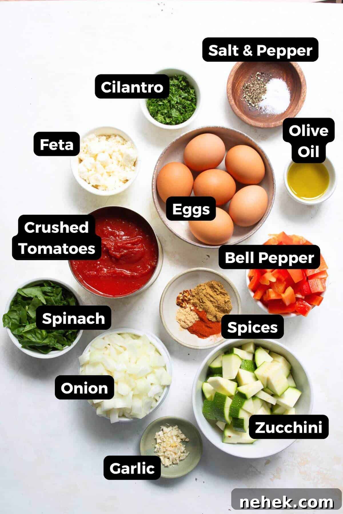 All ingredients needed to make Mediterranean shakshuka with feta, labeled and ready to cook-including eggs, zucchini, red bell pepper, yellow onion, garlic, spinach, cilantro, crushed tomatoes, cumin, paprika, ground ginger, red chili flakes, olive oil, and crumbled feta.