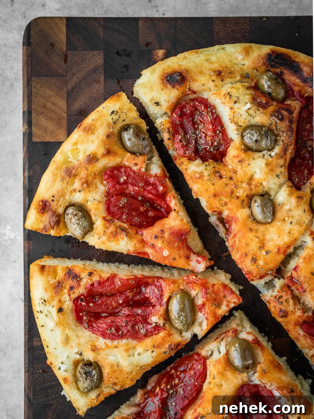 Focaccia Barese slices garnished with tomatoes, green olives and oregano on a wooden board.