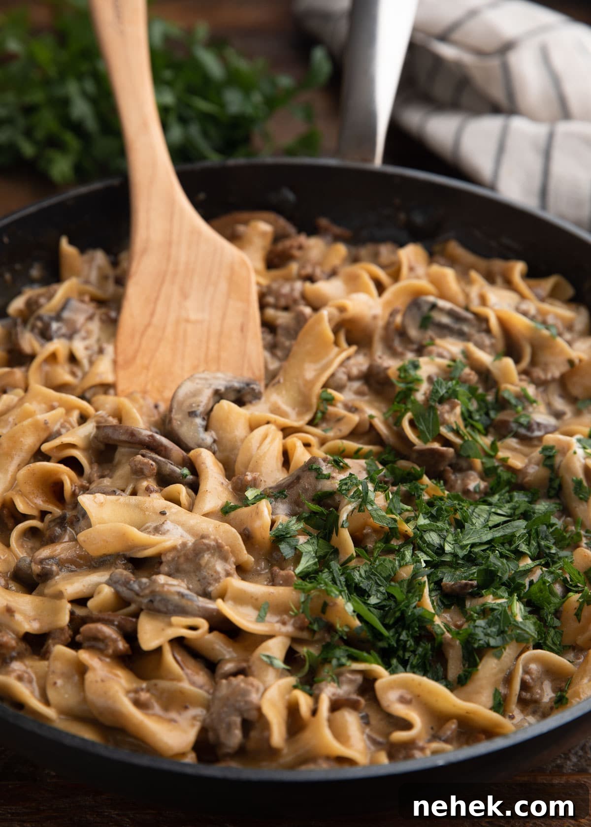 Skillet filled with creamy ground beef stroganoff, topped with a generous handful of chopped parsley and stirred with a wooden spoon.