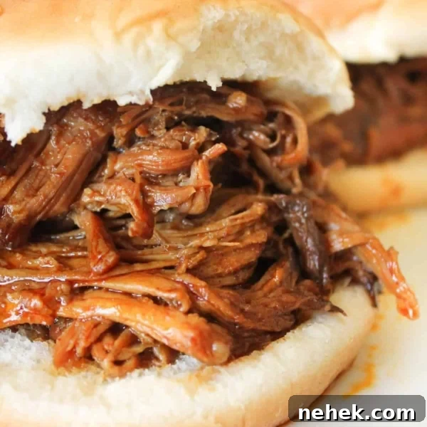 A close-up of a sandwich with shredded barbecue meat filling on a white bun, with another similar sandwich in the background.