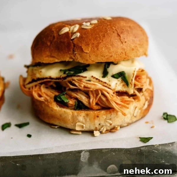 A sandwich with shredded chicken, melted cheese, and greens in a sesame seed bun, placed on white parchment paper.