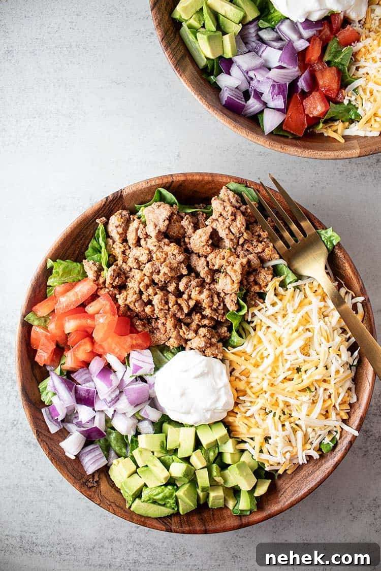 overhead view of turkey taco salad in a wooden bowl with sections of ground turkey, cheese, chopped avocado, chopped tomato, chopped onions and sour cream in the middle, all on a bed of lettuce and an additional bowl further toward the top