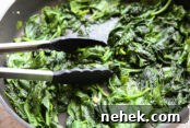 A horizontal photo of a large frying pan full of cooked spinach with a pair of tongs partially in the left side of the pan.