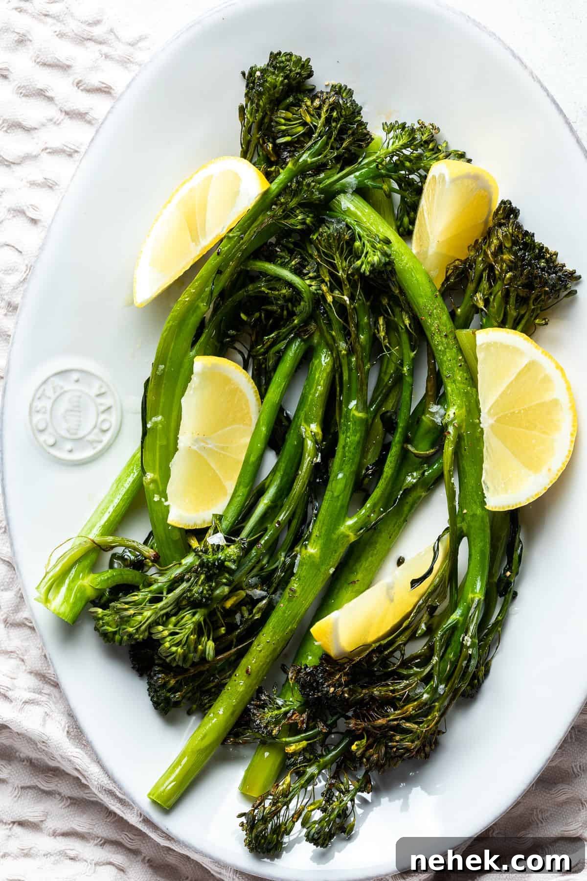 White dish with cooked broccolini garnished with lemon wedges.