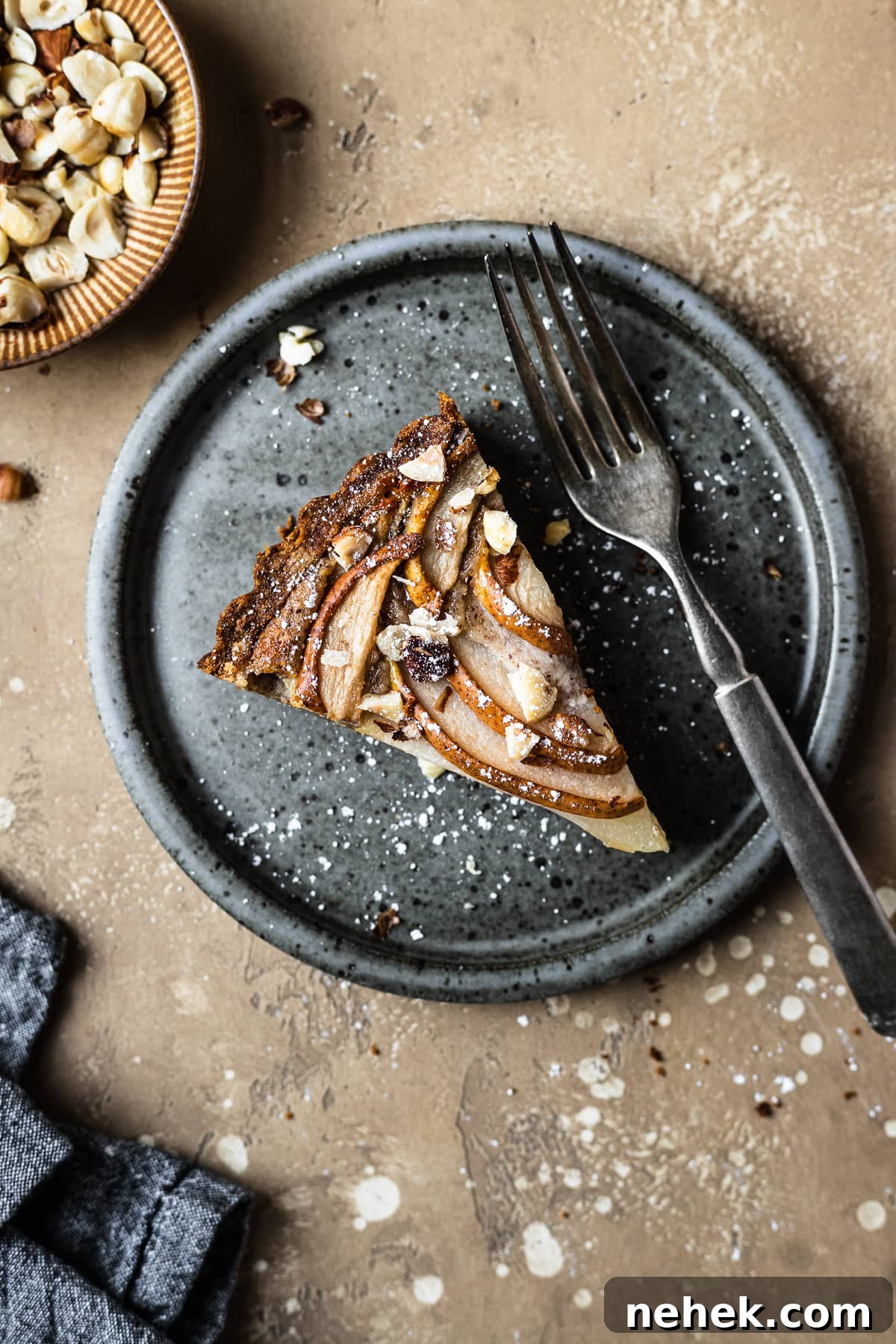 Pear Tart with Hazelnut Frangipane Recipe for a Crispy, Nutty Dessert 2 Pear hazelnut tart slice on a speckled grey blue ceramic plate. A vintage fork rests on the right side of the plate. The plate is on a warm tan textured stone background. A small bit of a textured grey napkin peeks out from the bottom left corner, and a small view of a bowl of chopped hazelnuts is visible at top left.