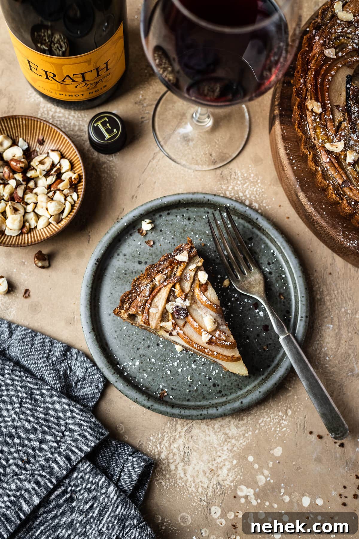 Pear Tart with Hazelnut Frangipane Recipe for a Crispy, Nutty Dessert 12 Pear hazelnut tart slice on a speckled grey blue ceramic plate. A vintage fork rests on the right side of the plate. The plate is on a warm tan textured stone background. A textured grey napkin is at the bottom left corner, and a small bowl of chopped hazelnuts is visible at top left along with a wine bottle and a glass of red wine at top. The remainder of the tart peeks out at top right.