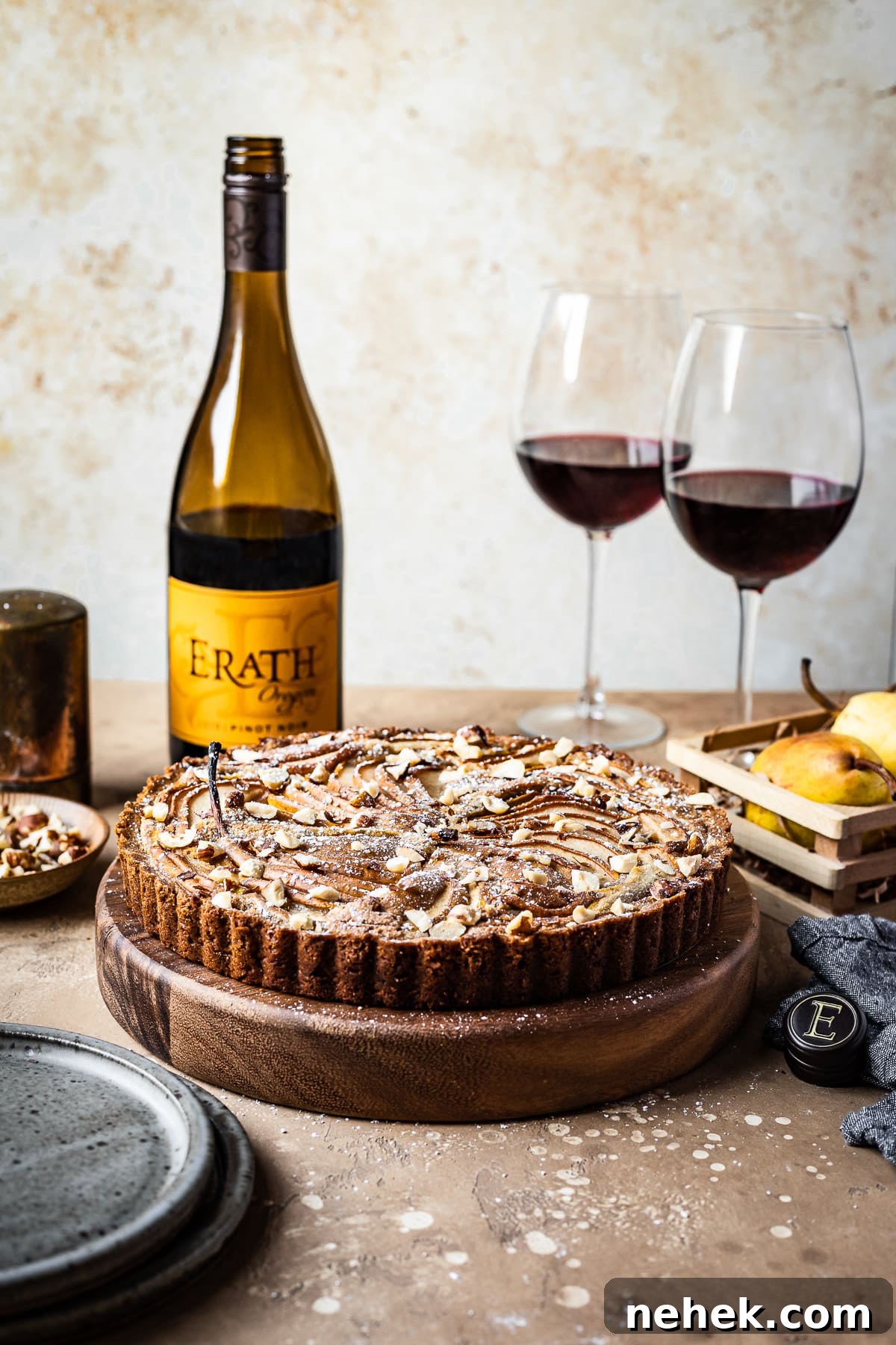 Pear Tart with Hazelnut Frangipane Recipe for a Crispy, Nutty Dessert 3 A front view of a pear tart on a round wooden platter on a textured warm tan background. The tart is surrounded by blue grey ceramic plates, an opened bottle of red wine, two half full glasses of red wine, a small wooden crate of pears, and a textured grey napkin.