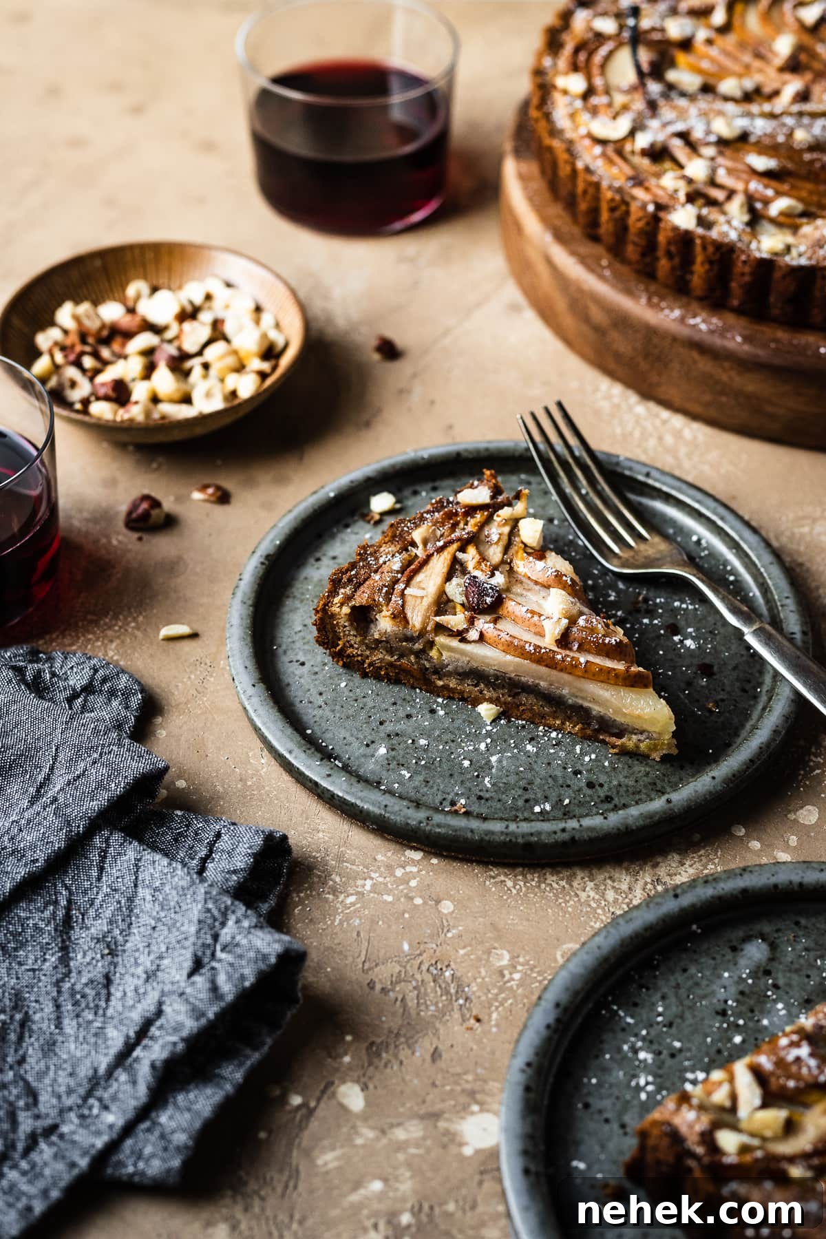 Pear Tart with Hazelnut Frangipane Recipe for a Crispy, Nutty Dessert 4 An angled view of two slices of tart on speckled grey blue ceramic plates. Nearby is the remainder of the tart, a small bowl of hazelnuts, and two stemless wine glasses filled with red wine. A grey textured linen napkin is at bottom left. The background of the image is a warm tan speckled stone.