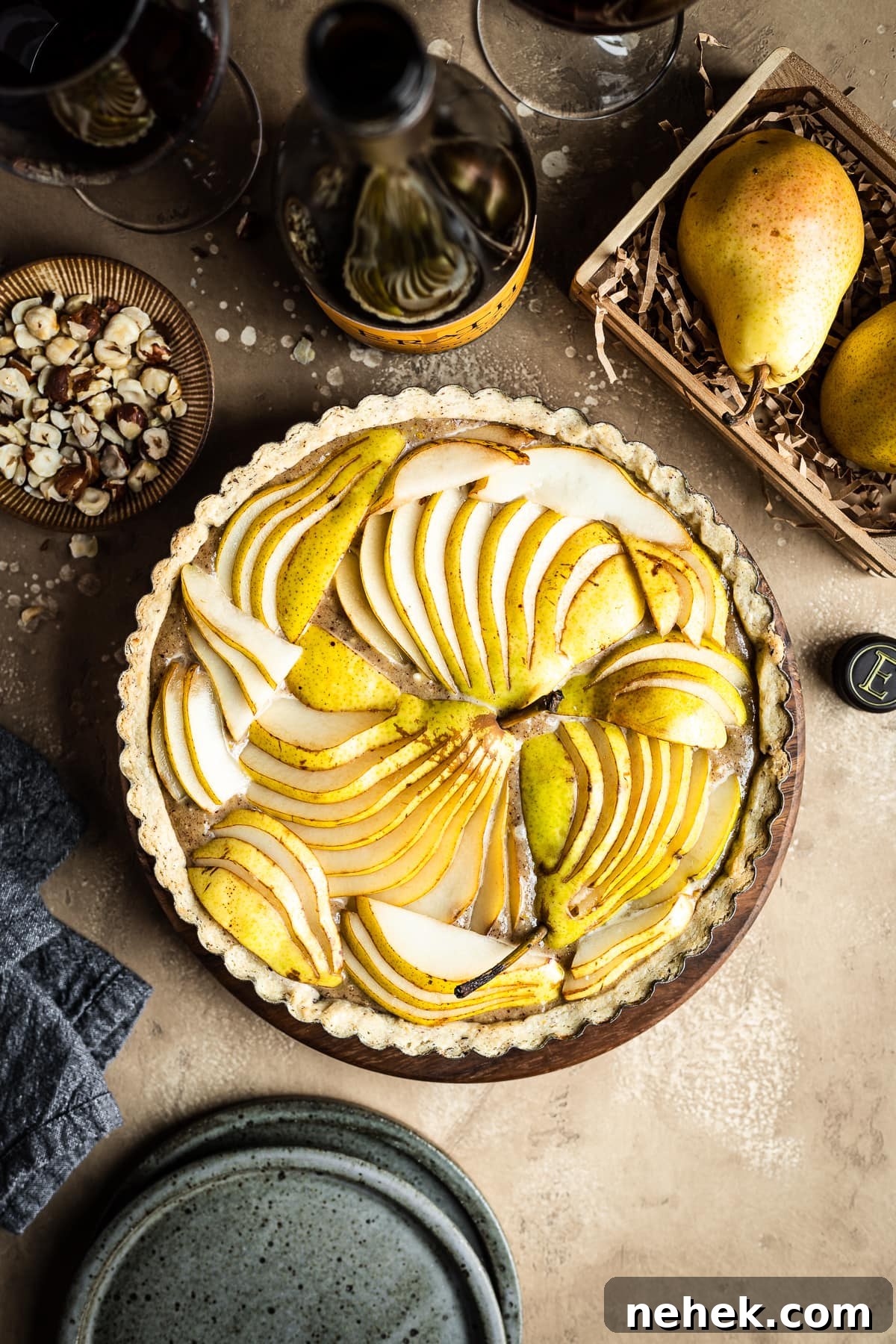 Pear Tart with Hazelnut Frangipane Recipe for a Crispy, Nutty Dessert 5 An unbaked pear hazelnut tart in a round pan is at the center of the image. Surrounding the tart are speckled blue ceramic plates, a bowl of chopped hazelnuts, pears in a wooden crate, a linen napkin, and a bottle of red wine with two filled wine glasses. Everything rests on a textured tan background.
