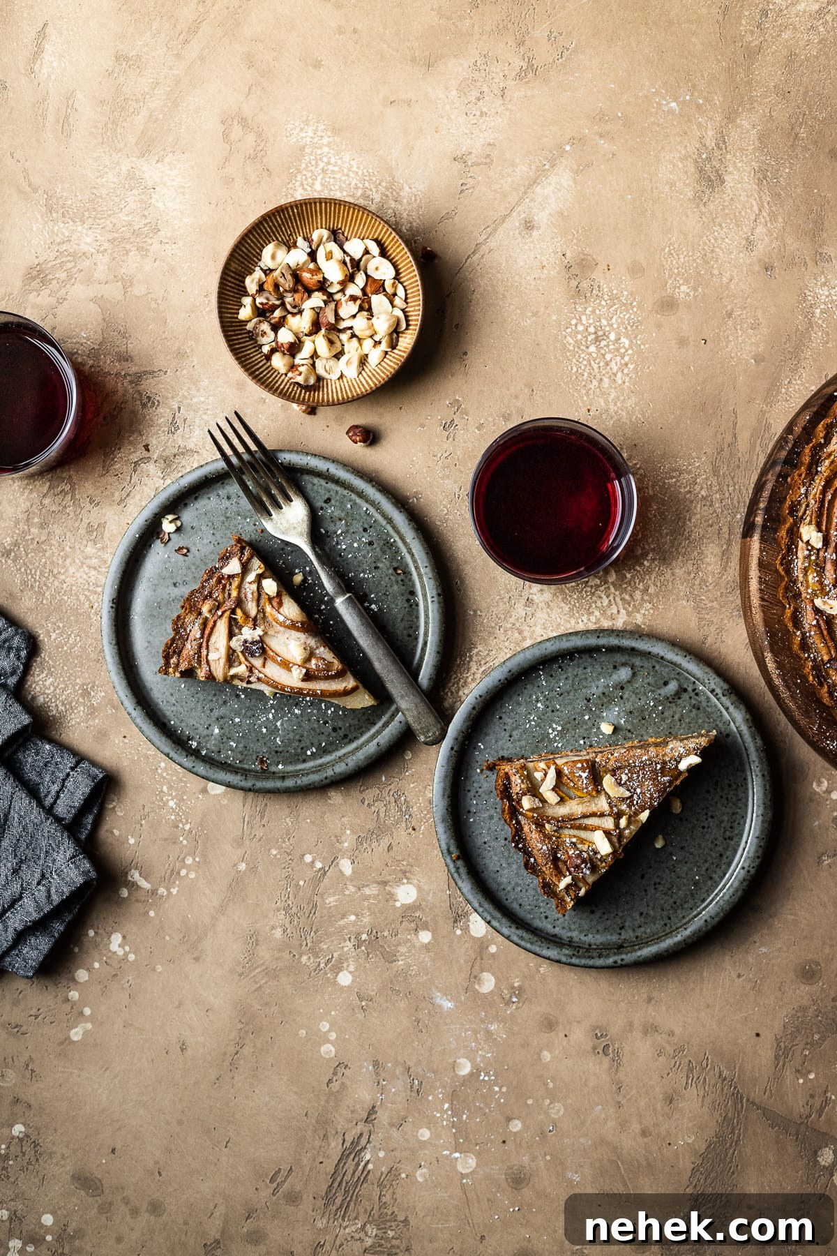 Pear Tart with Hazelnut Frangipane Recipe for a Crispy, Nutty Dessert 7 Two slices of tart on speckled grey blue ceramic plates. To the right is the remainder of the tart. A small bowl of hazelnuts, and two stemless wine glasses filled with red wine surround the plates. A grey textured linen napkin is at lower left. The background of the image is a warm tan speckled stone.