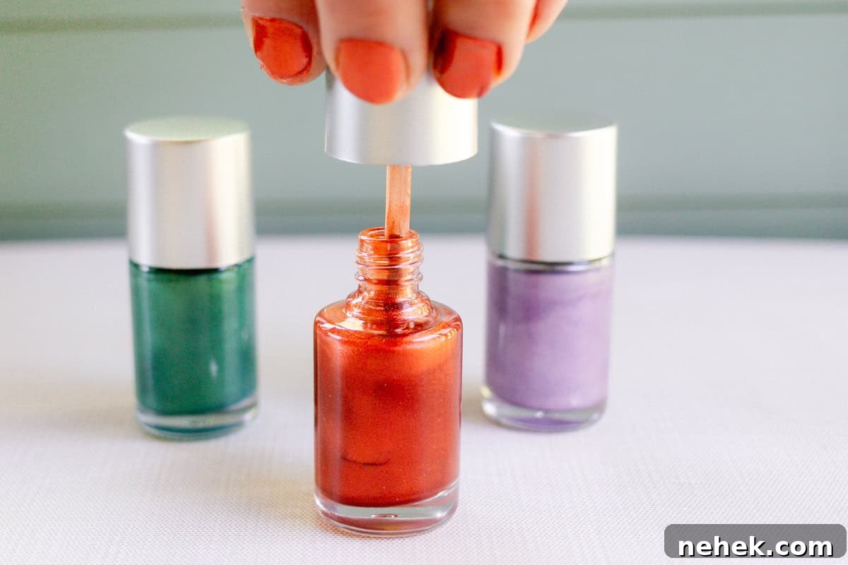 A hand with red nails pulling the brush out of a bottle of homemade red nail polish. A green bottle and a purple bottle of homemade nail polish are in the background.