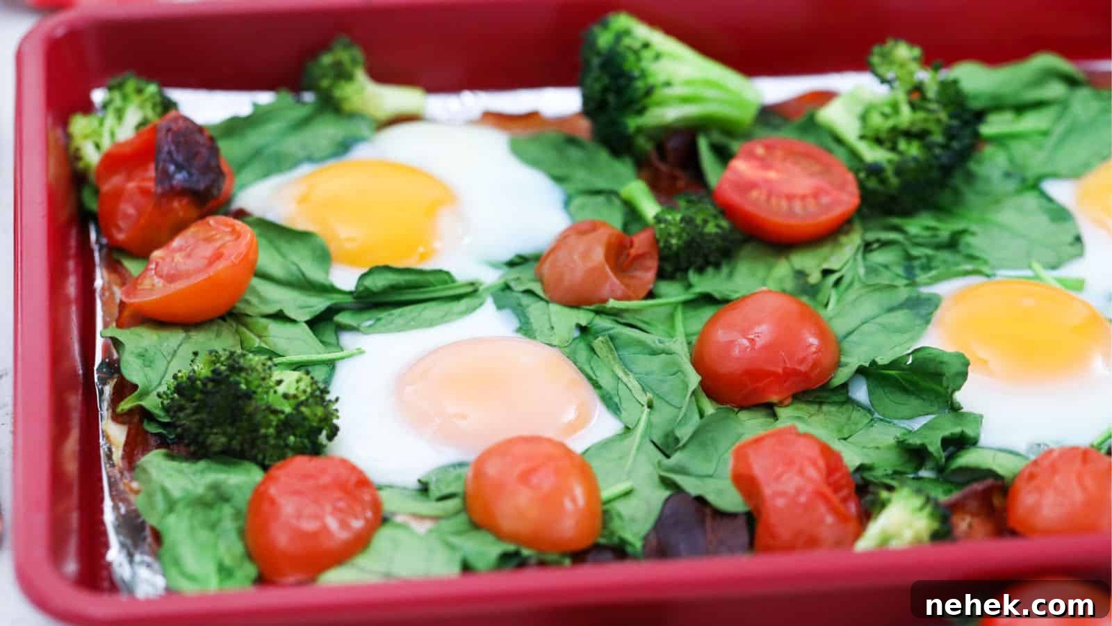 Baked eggs with spinach, broccoli, and cherry tomatoes in a red baking dish.