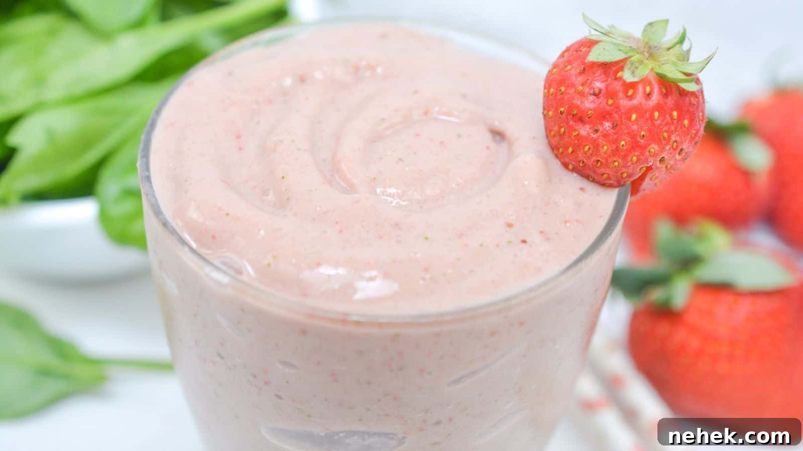 A glass of strawberry smoothie garnished with a whole strawberry, surrounded by fresh strawberries and spinach leaves.