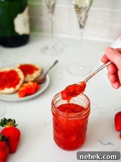 A hand scoops strawberry champagne jam out of a jar. Two champagne glasses stand behind it.