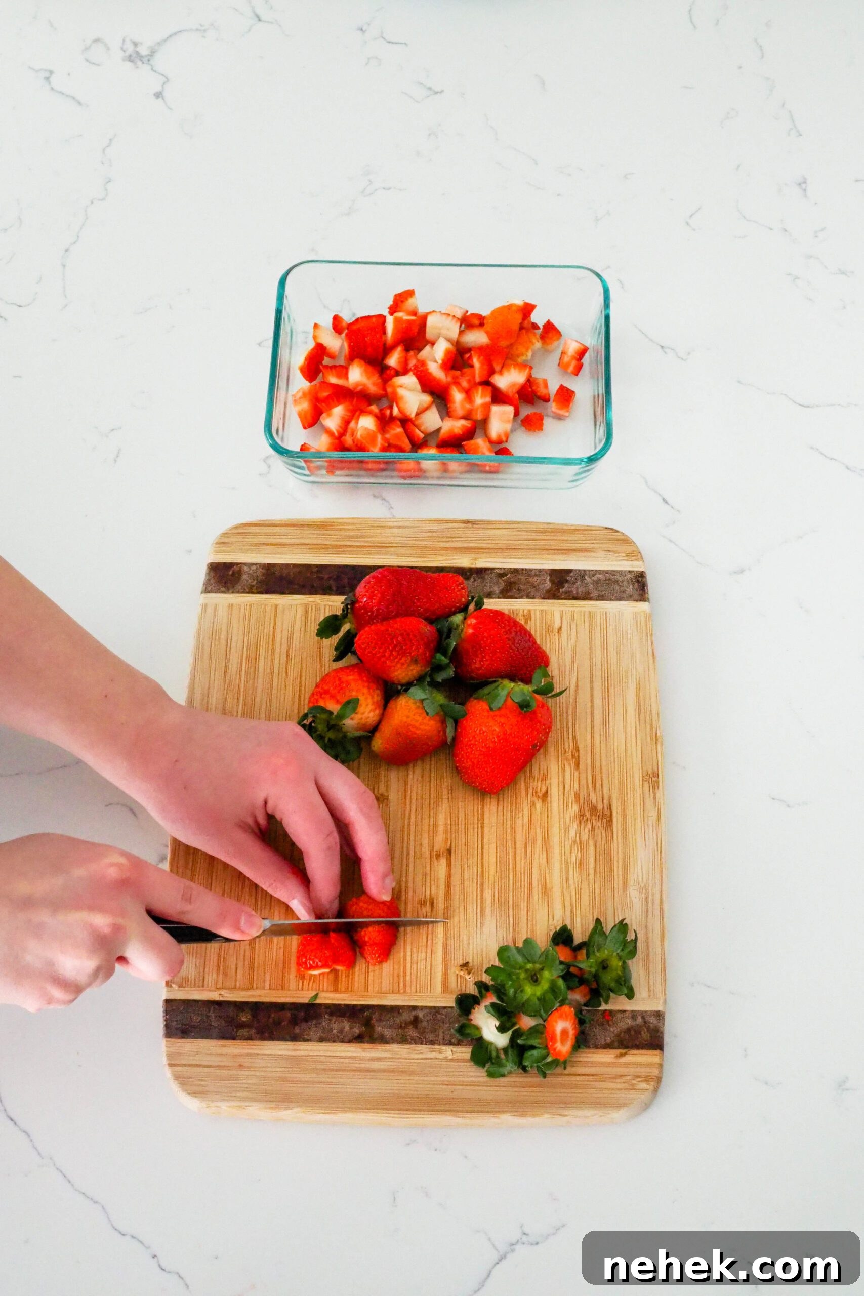 Two hands dice strawberries on a wooden cutting board.