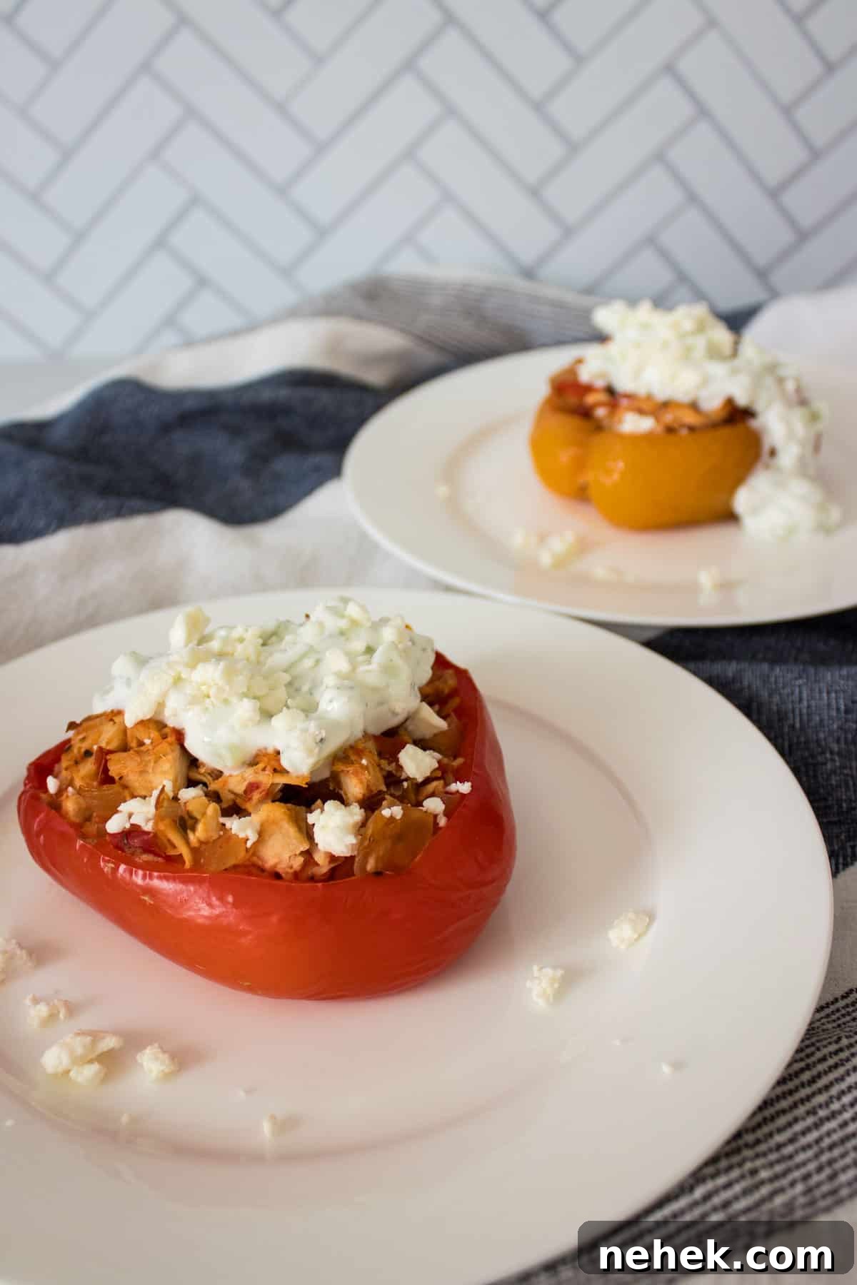 Two plates with a stuffed bell pepper half slathered with tzatziki and garnished with feta on a blue and white towel with one plate in the front and the other in the background.