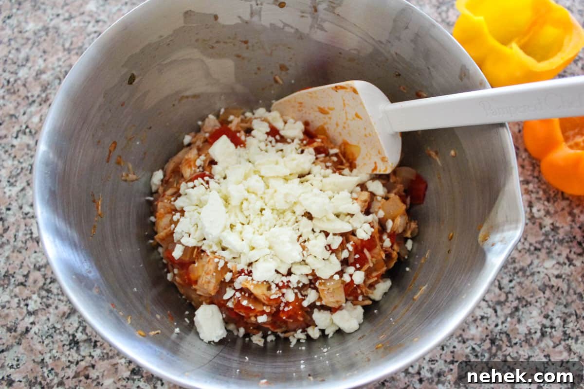 Vegetables and spices mixed in a stainless bowl with feta cheese on top about to be mixed in, and bell peppers to the side of the bowl.