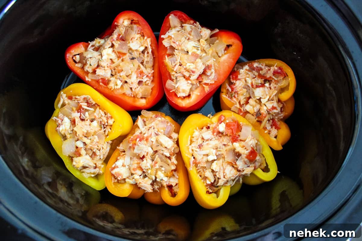 Six bell pepper halves stuffed with a mixture of chicken, cheese, and vegetables, arranged in an oval slow cooker.