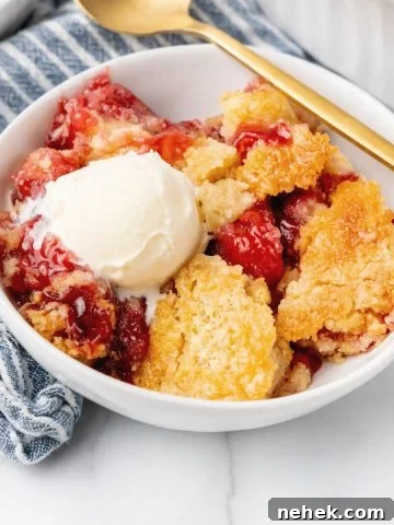 Striped blue white kitchen towel with white bowl containing serving of cherry cobbler with ice cream. A gold spoon, white marble surface.