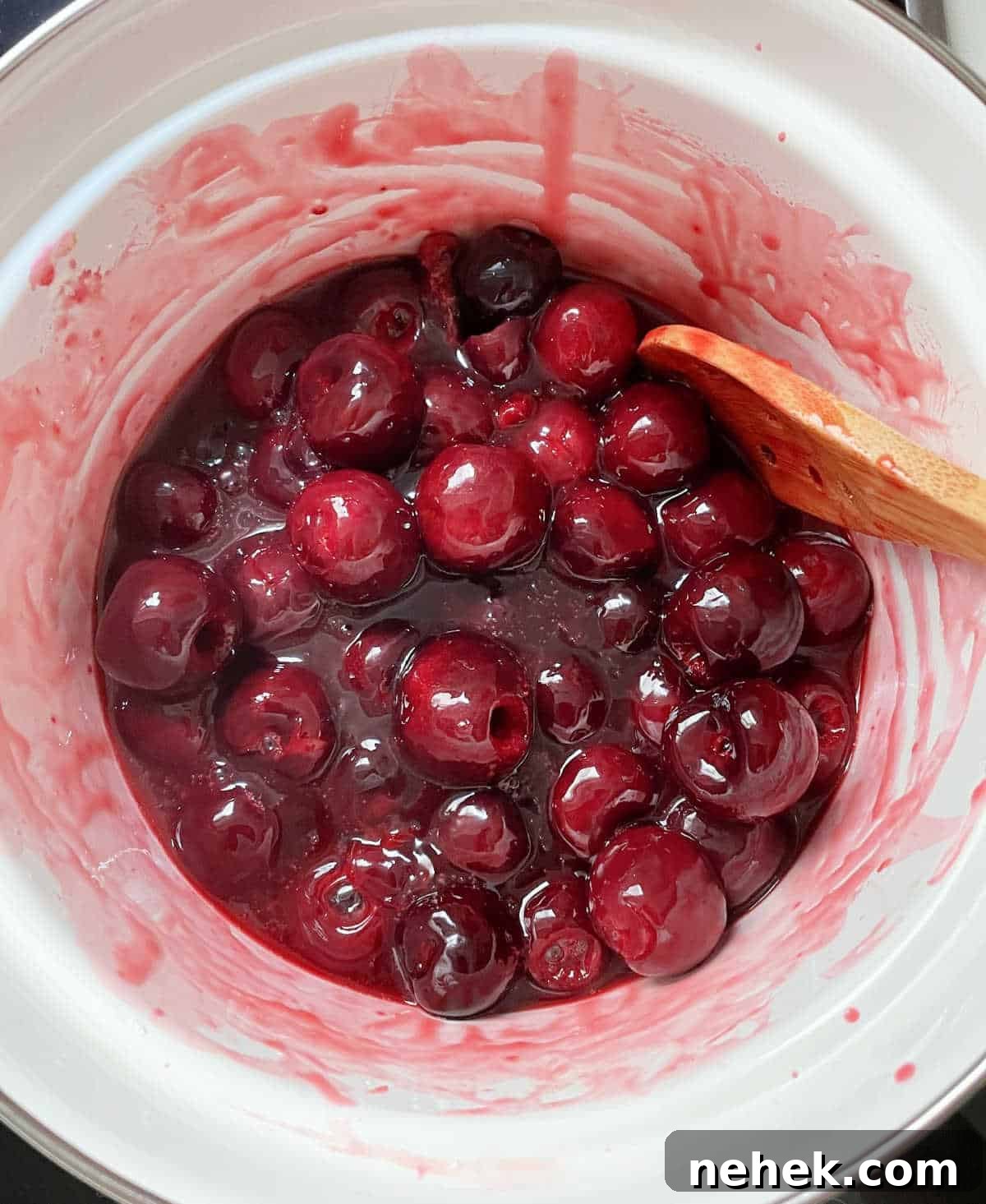 Top view of cherry pie filling simmering in white saucepan with wooden spoon.