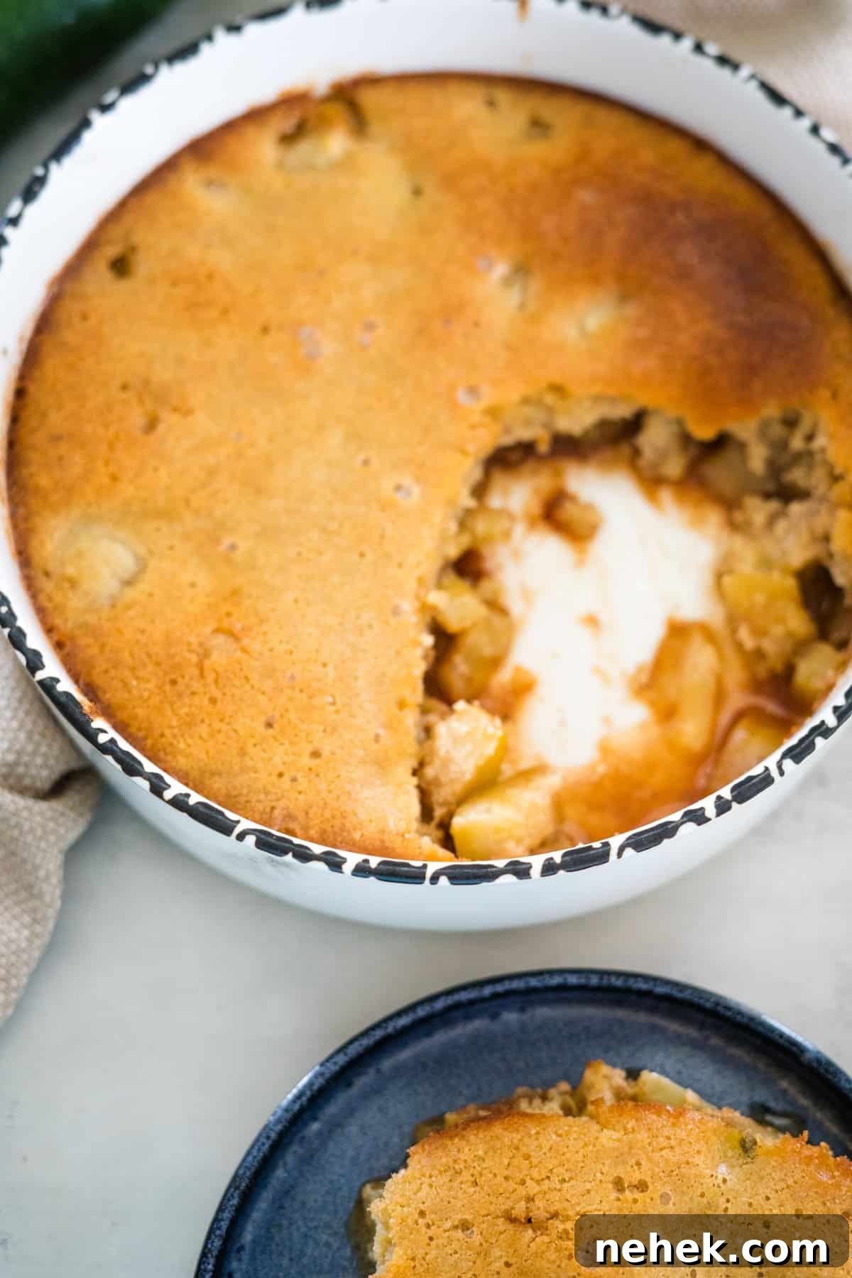 A baked apple cobbler in a white round dish with a portion scooped out, revealing the filling. A plate with a serving of the cobbler sits next to the dish.
